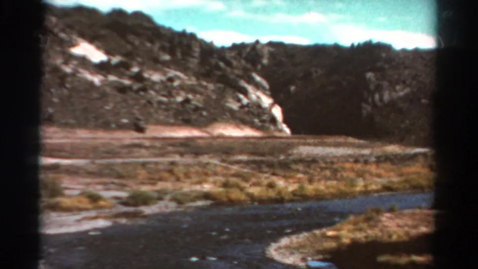 1967 Colorado home-movie frame showing a curving river, gravel bank, dry grasses, and steep rocky hills in daylight.