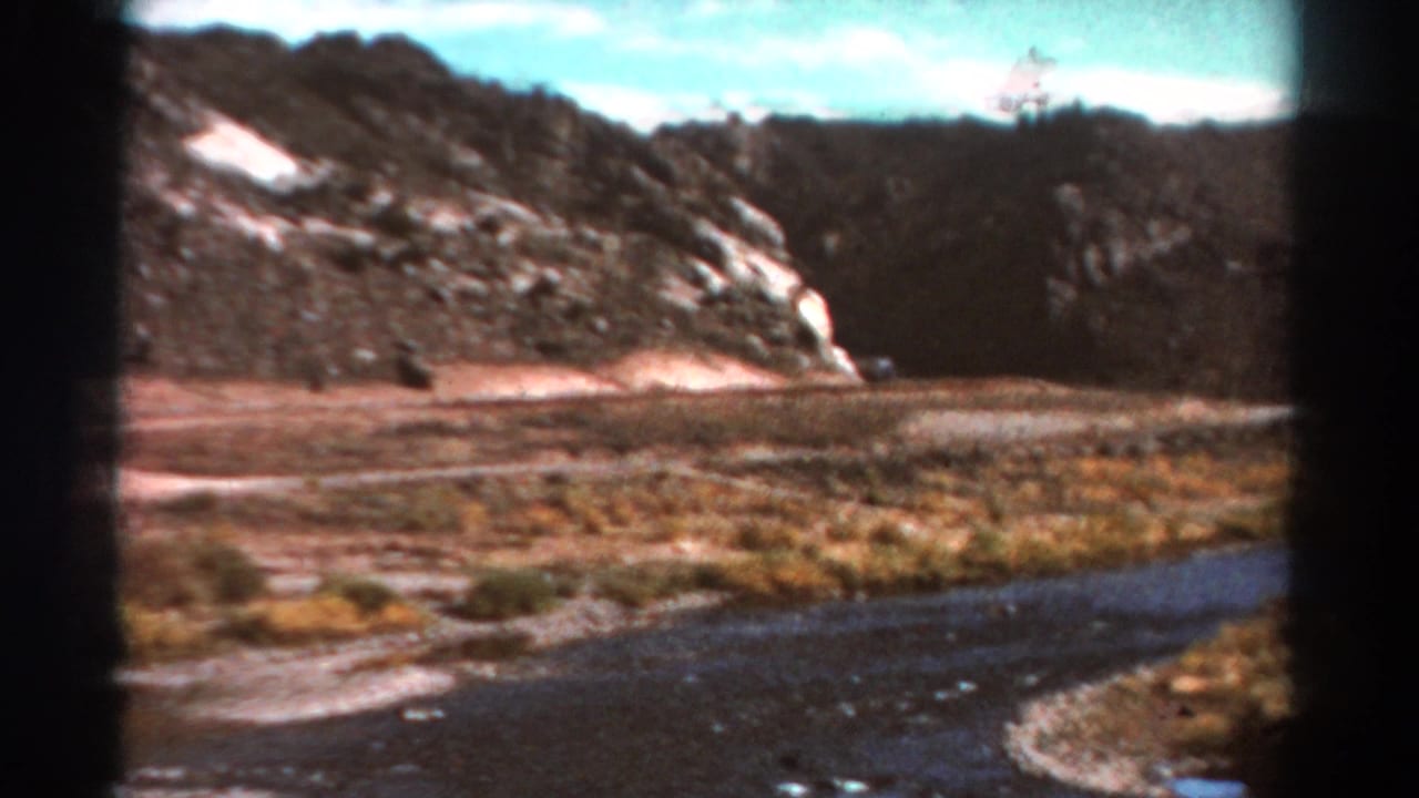 River foreground with dry valley floor, rocky hillside, dark tree line, and pale clouds.