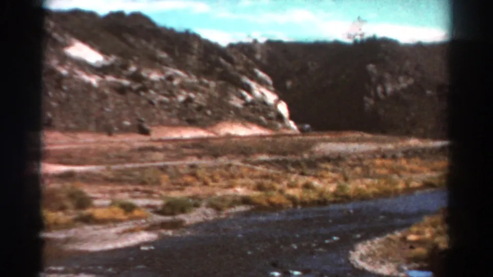 1967 Colorado home-movie frame with a river in the foreground, dry valley grasses, rocky hillside, and dark evergreen ridge.