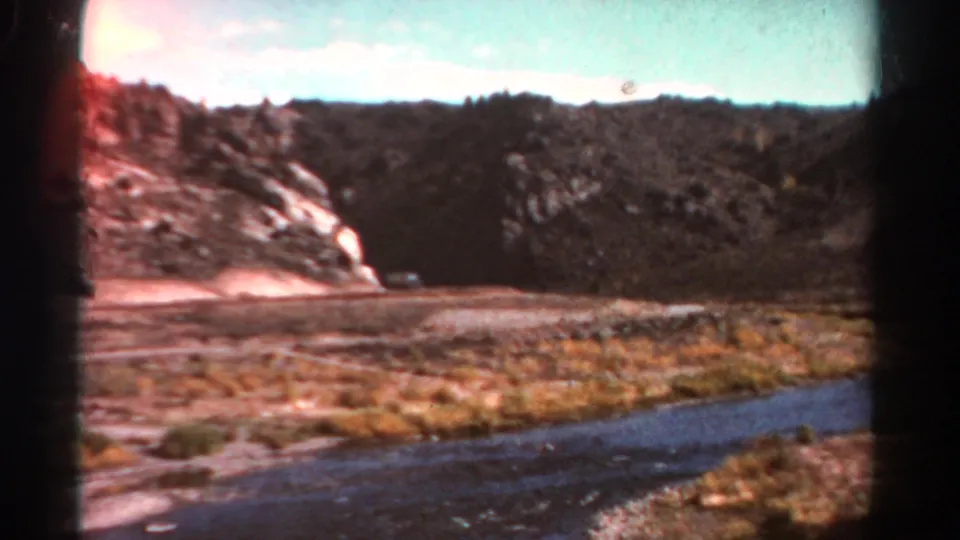 1967 Colorado 8mm frame showing a wider look across a dry mountain valley, with river water, scrub grass, rocky slope, and distant hills.