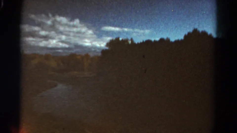 1967 Colorado 8mm frame with a darker wooded slope, faint river or path line, low hills, blue sky, and scattered clouds.