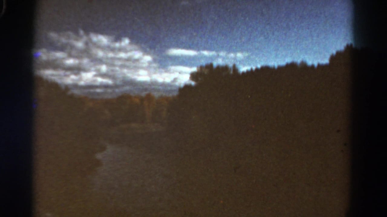 1967 Colorado 8mm frame showing a river or creek curving past dense trees beneath scattered clouds and blue sky.