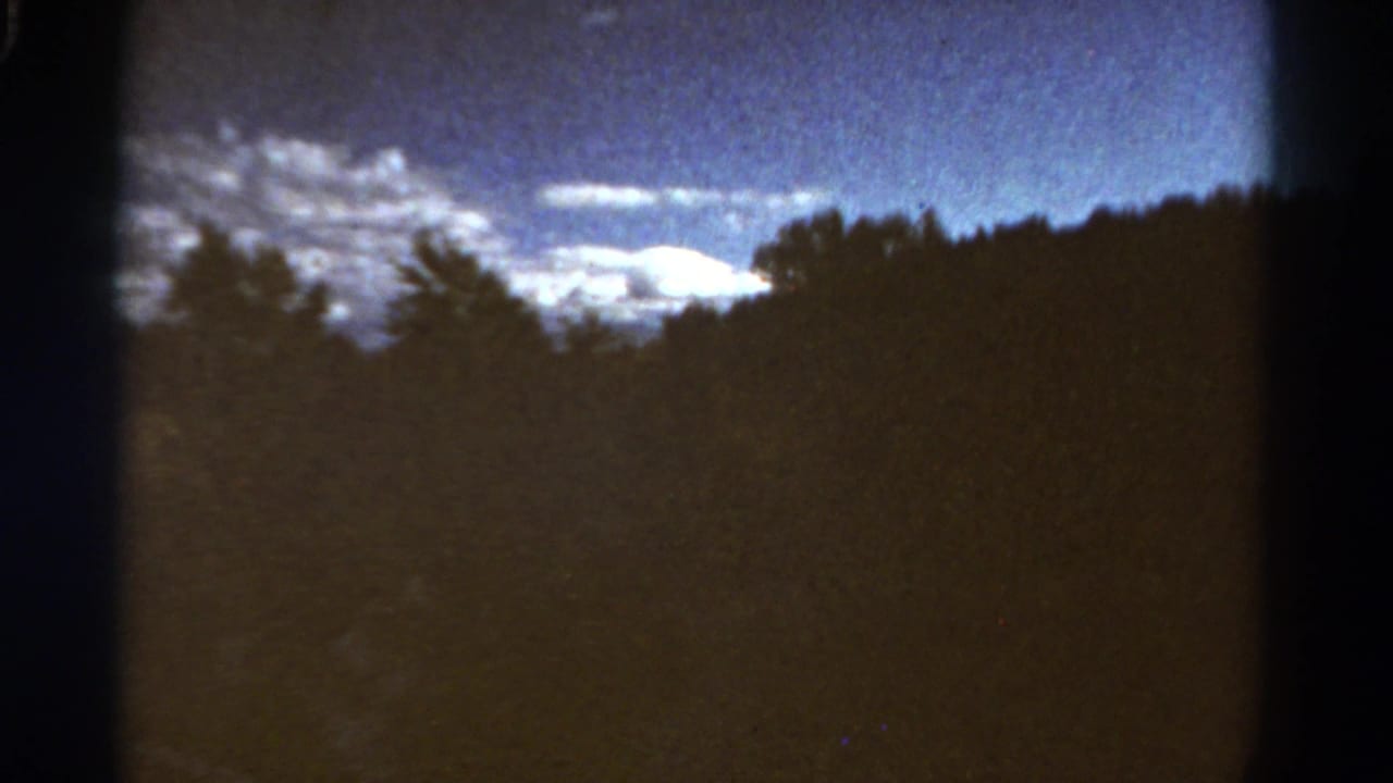 1967 Colorado home-movie frame with dense trees in silhouette below a bright cloud band and blue sky.