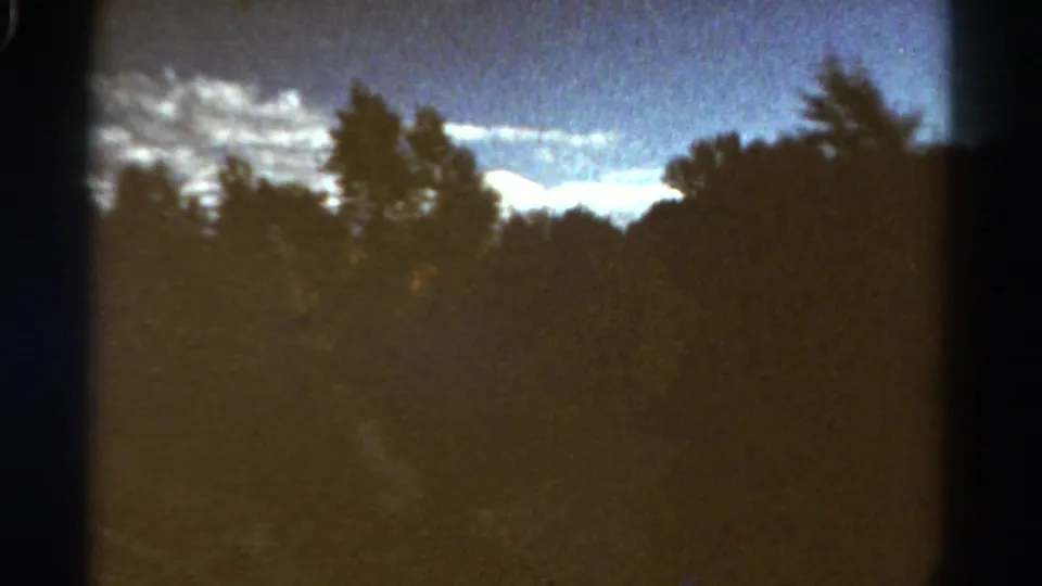 1967 Colorado 8mm frame showing blurred roadside trees, a possible river or track, and bright clouds over the forest.