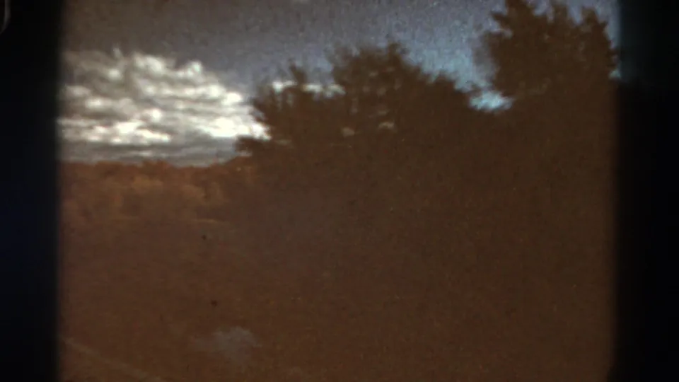1967 Colorado home-movie frame with dark trees, a wooded slope, and broken clouds above the landscape.