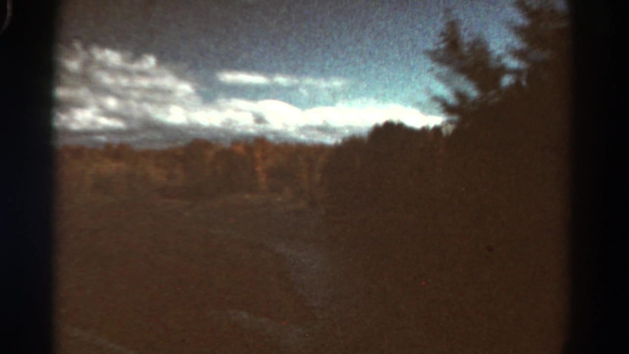 1967 Colorado 8mm frame showing an open field, tree line, distant ridge or mountains, and scattered clouds under a blue sky.