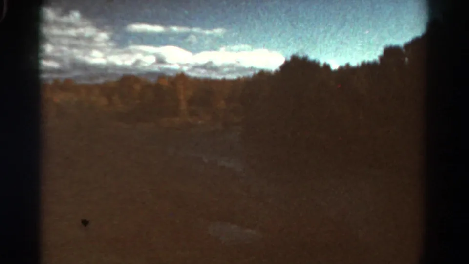 1967 Colorado home-movie frame with a shadowed field, dense trees at right, distant landforms, and bright layered clouds.