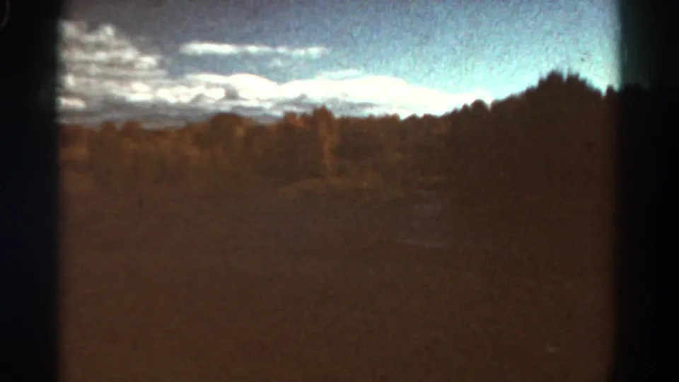 1967 Colorado home-movie frame with a tree-lined field, distant ridge or mountains, bright clouds, blue sky, and dark frame edges.