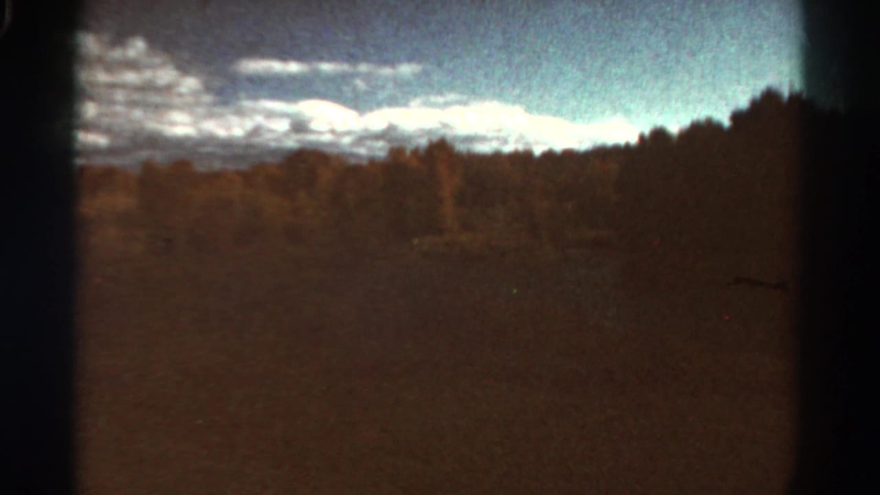 1967 Colorado home-movie frame showing distant snow-covered mountains, a dark treeline, and open countryside seen through heavy 8mm grain.