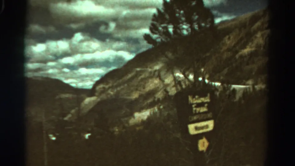 1967 Colorado 8mm frame with a wooden National Forest sign, hillside, pine trees, mountains, and cloudy sky.