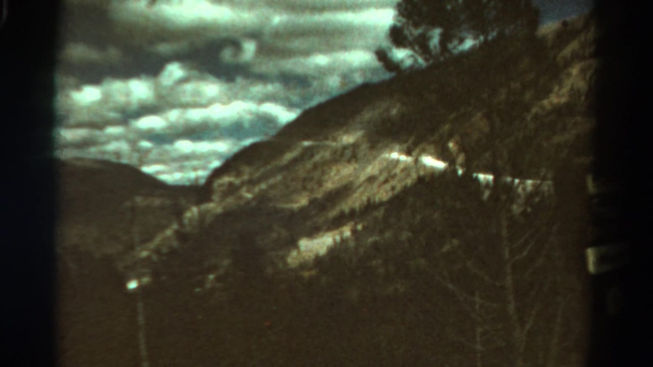 1967 Colorado 8mm home-movie frame showing a mountain slope, road cut, cloudy sky, and dark valley foreground.