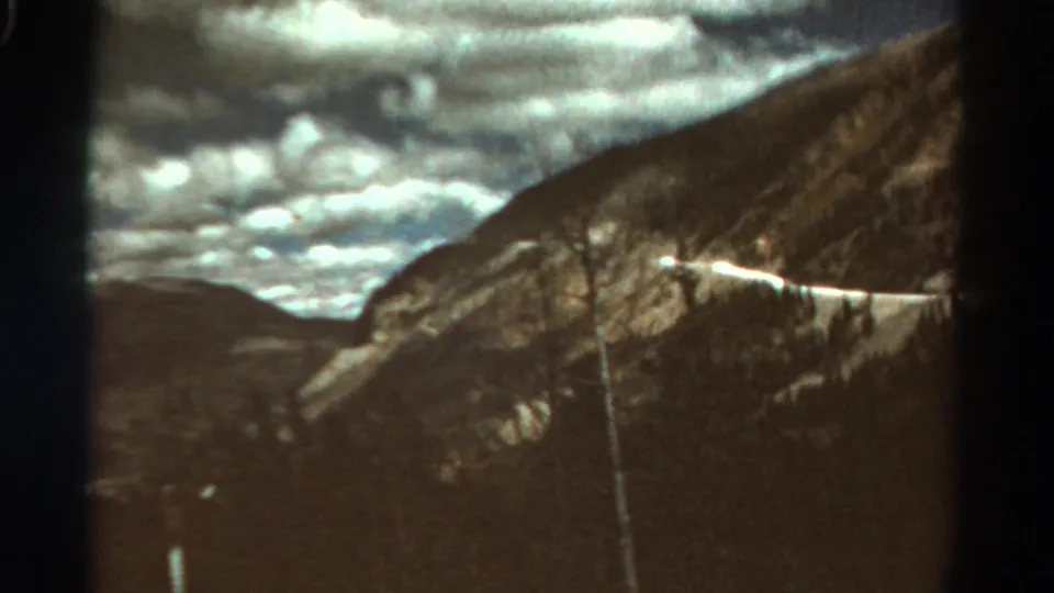 1967 Colorado home-movie frame showing the road cut along a mountain slope with dark foreground and cloudy sky.