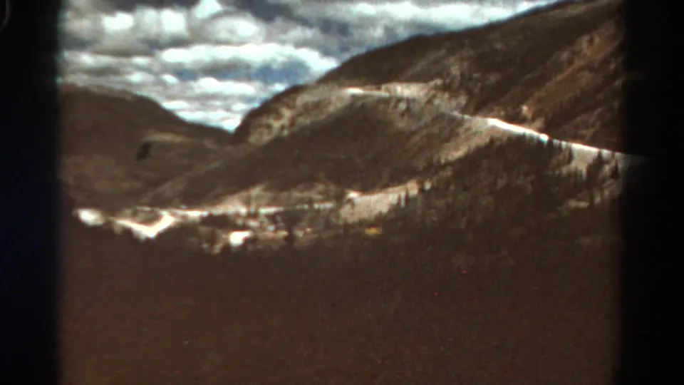 1967 Colorado 8mm frame showing a wide mountain valley, road cut, pale snow or rock patches, and heavy clouds.