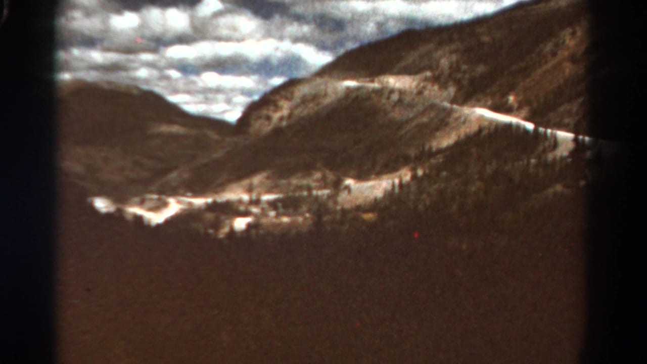 1967 Colorado 8mm frame showing a distant mountain valley, sunlit cloud cover, forested slopes, and pale snow or road traces.