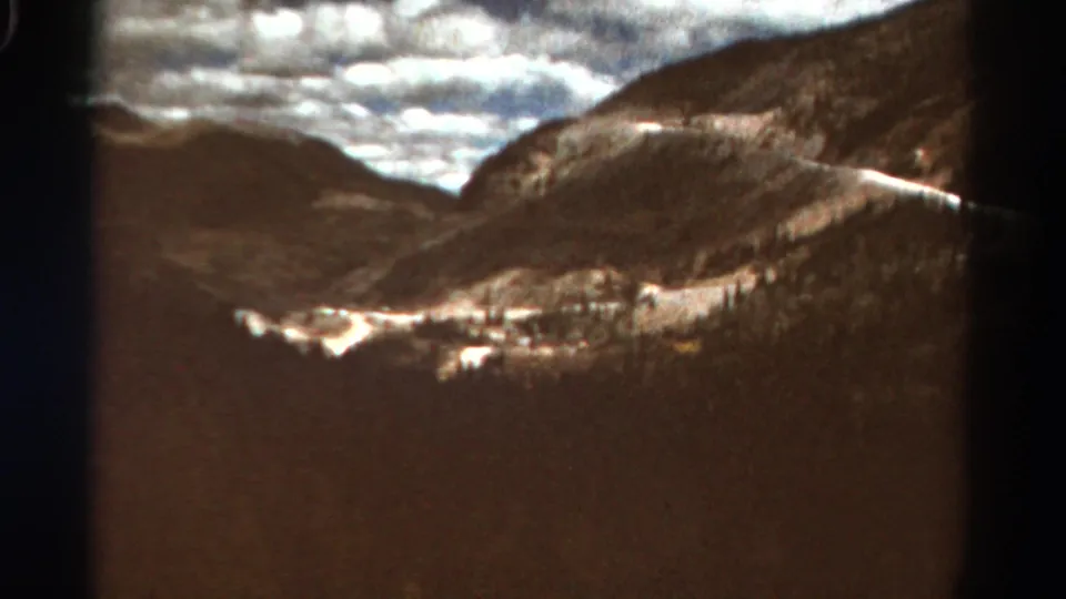 1967 Colorado home-movie frame showing wooded mountain slopes, a valley below, broken cloud light, and pale patches of snow or roadway.