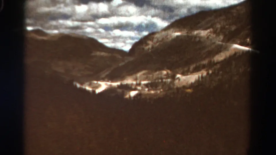 1967 Colorado 8mm frame with distant ridges, forested slopes, a valley road or snow trace, and bright clouds over the mountains.