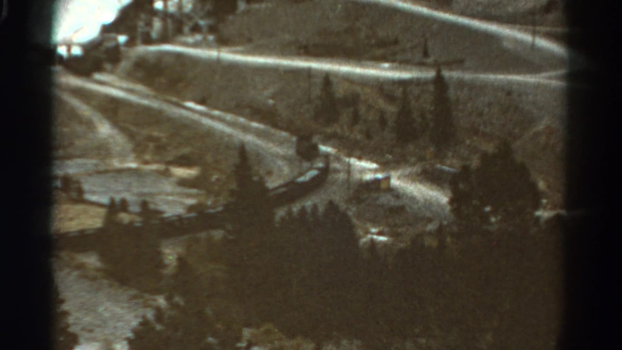 1967 Colorado home-movie frame showing a passenger train on a curving mountain railway among evergreens and rocky slopes.