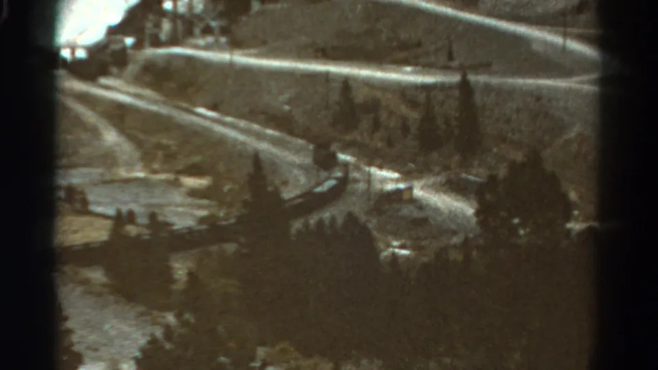 1967 Colorado home-movie frame showing a passenger train on a curving mountain railway among evergreens and rocky slopes.