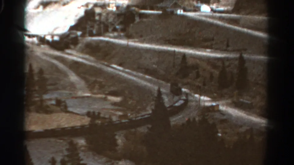 The train continues around the Colorado mountain curve, seen from above with evergreens, slopes, and visible 8mm frame borders.