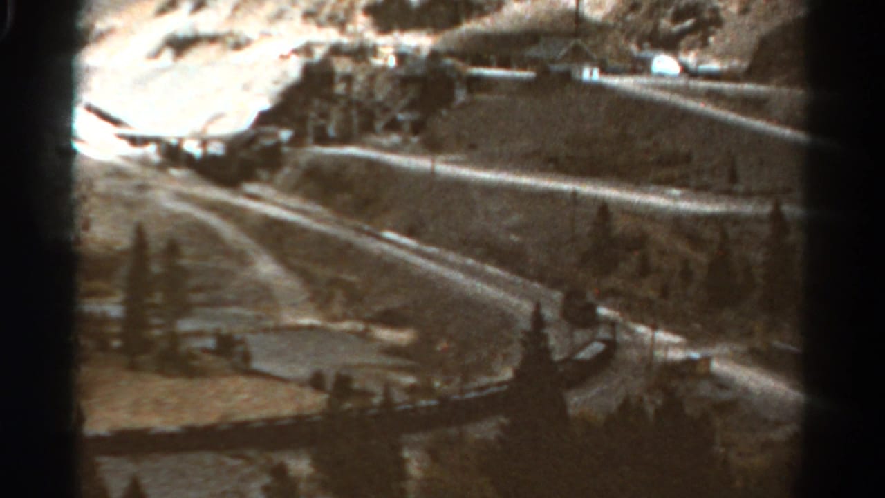 1967 Colorado 8mm frame showing a winding mountain road, hillside terrain, roadside edge, trees, and a small settlement in the distance.