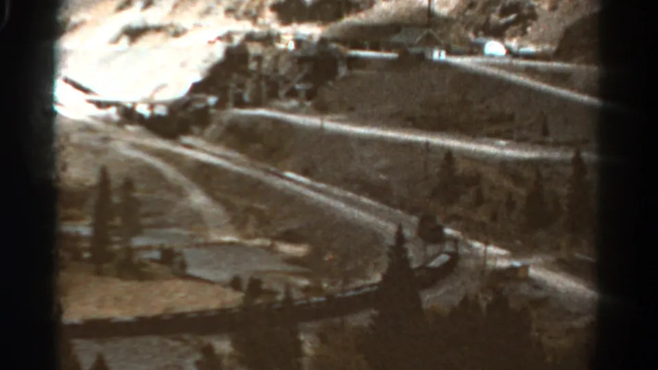 1967 Colorado 8mm frame showing a winding mountain road, hillside terrain, roadside edge, trees, and a small settlement in the distance.
