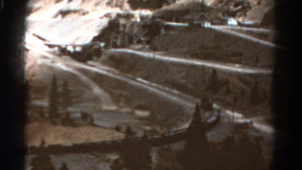 Winding road and roadside edge crossing a rugged landscape with trees and distant structures.
