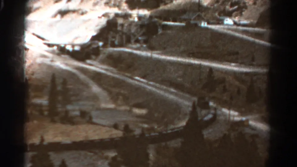 1967 Colorado home-movie view of a two-lane mountain road bending through a rugged, tree-dotted landscape with buildings beyond.