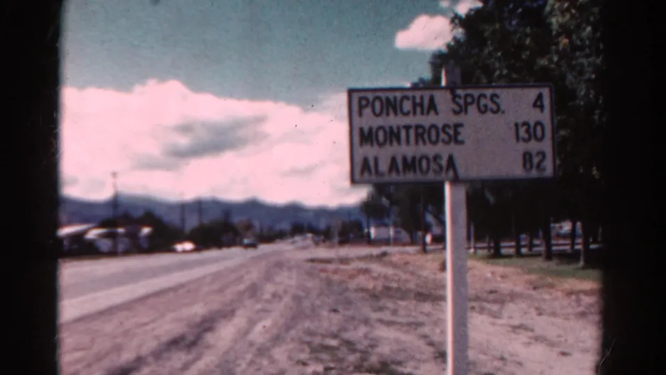 1967 Colorado 8mm frame with a roadside distance sign for Poncha Spgs., Montrose, and Alamosa beside a two-lane road and distant mountains.