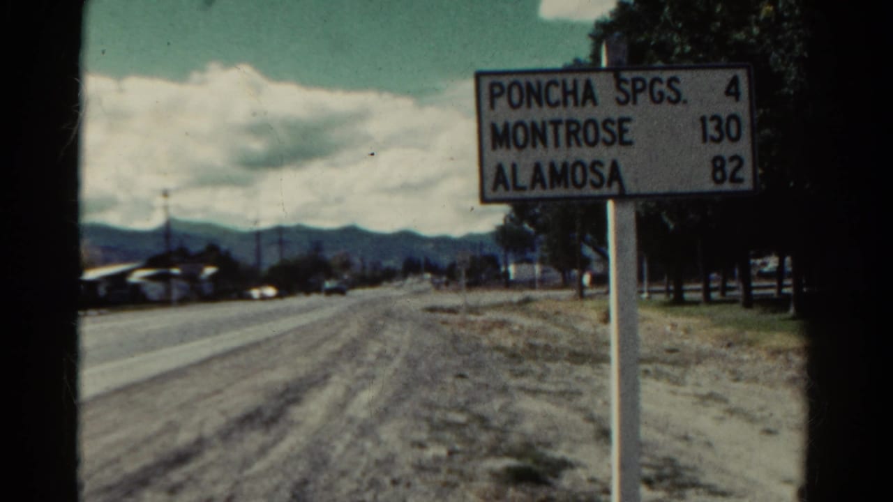 1967 Colorado home-movie frame with a roadside mileage sign and mountain horizon beyond the road.