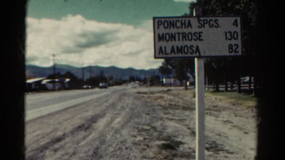 1967 Colorado travel frame with a roadside distance sign, rural road shoulder, trees, and distant mountains.