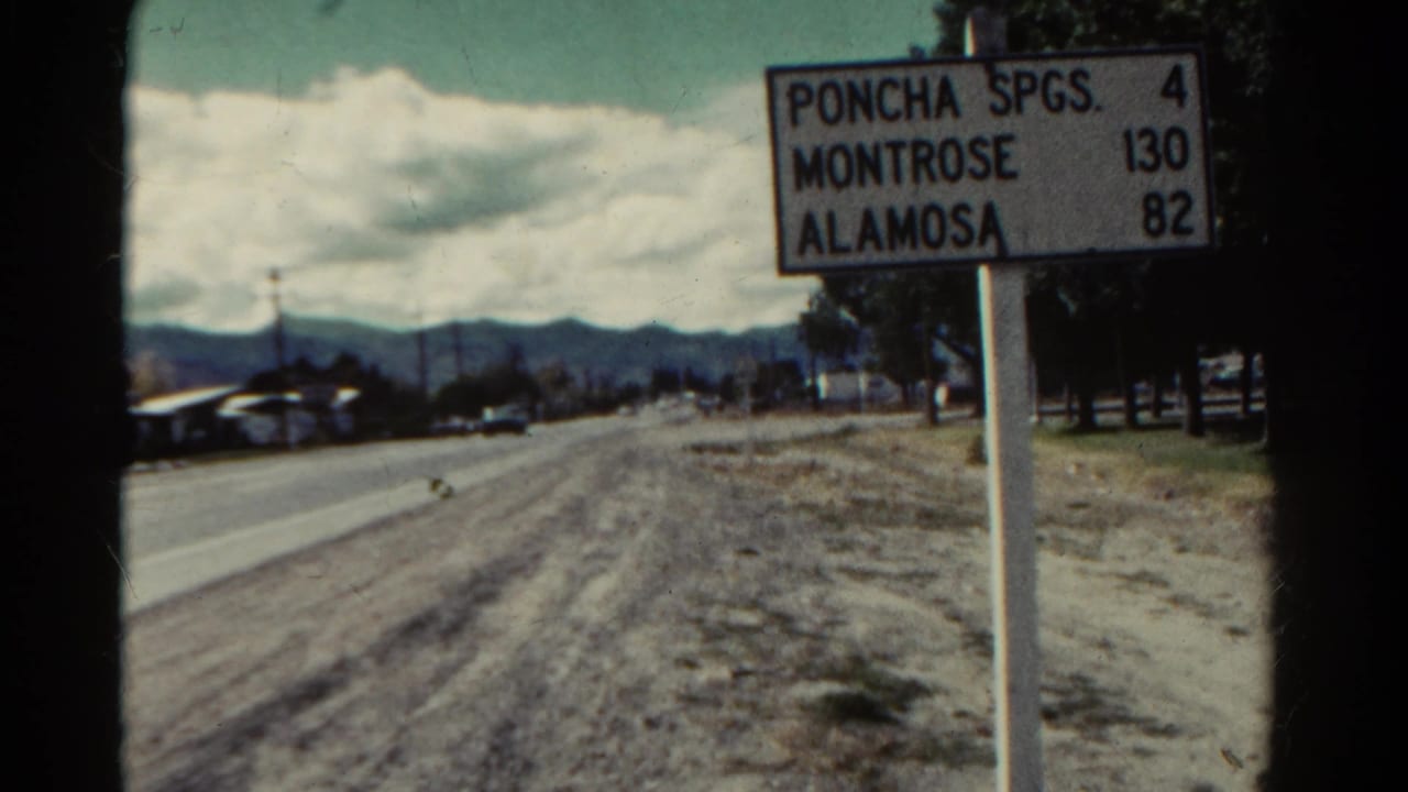 Closer roadside sign listing Poncha Spgs., Montrose and Alamosa near a road and mountains.