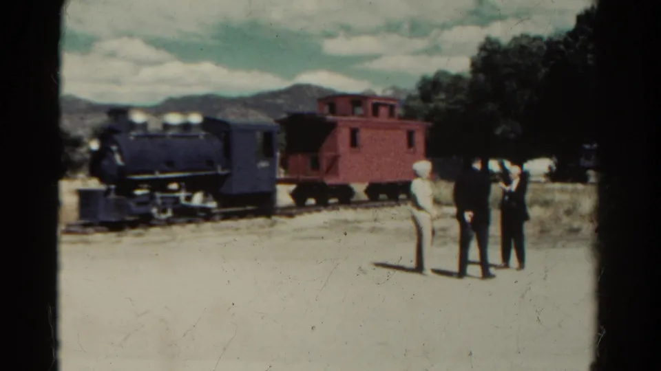 1967 Colorado home-movie frame showing three people observing a small black locomotive and red caboose in an open area.