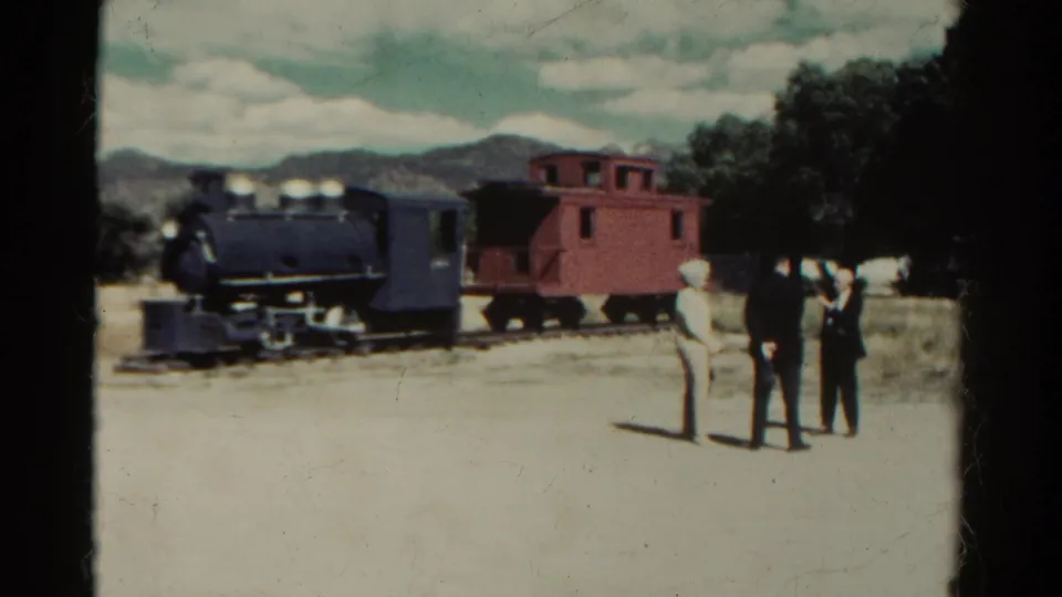 1967 Colorado frame of onlookers near a small steam train and caboose, with trees and mountains in the background.