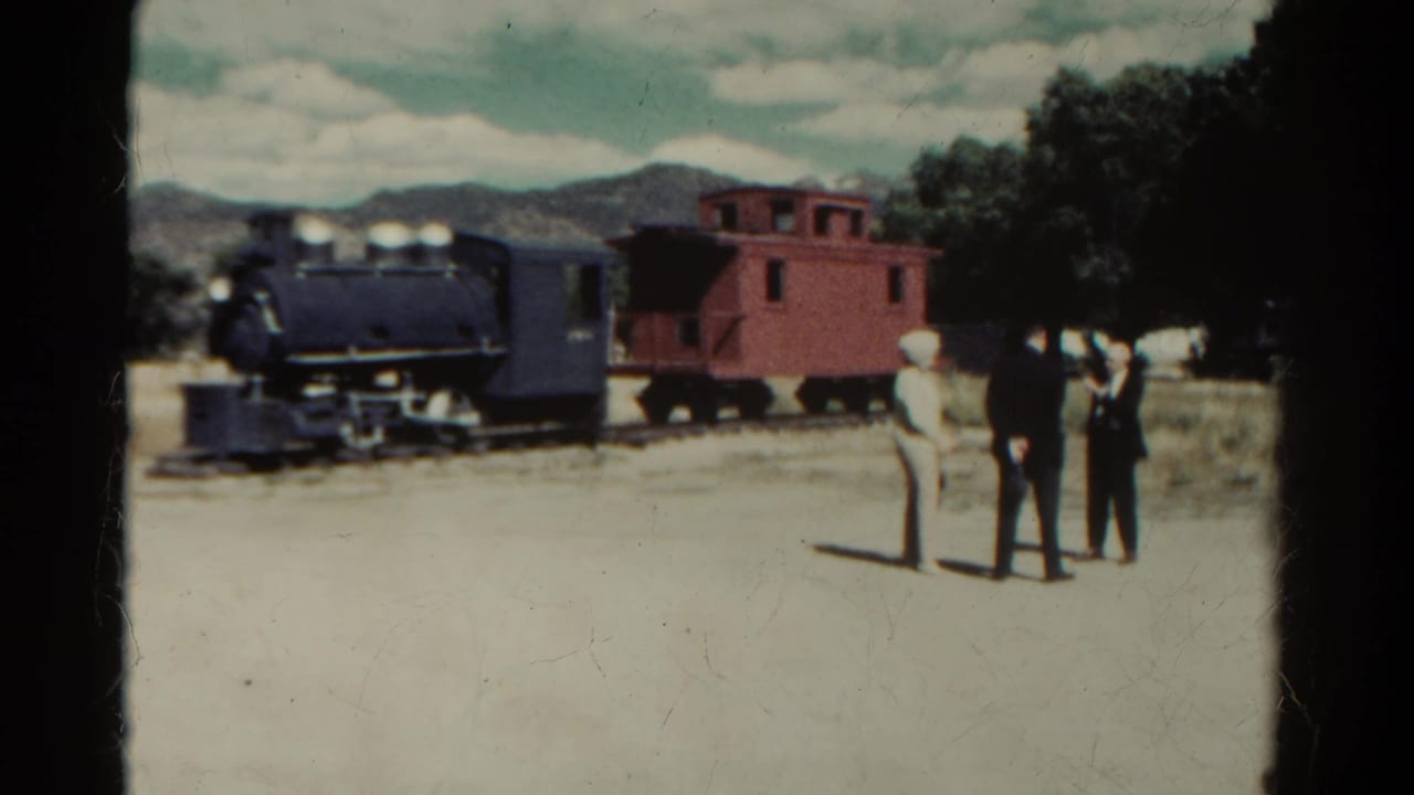 1967 Colorado home-movie frame showing a dark steam locomotive, red caboose, and three visitors on a dirt path with mountains behind.