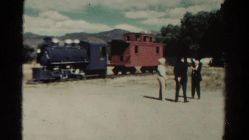 1967 Colorado home-movie frame showing a dark steam locomotive, red caboose, and three visitors on a dirt path with mountains behind.