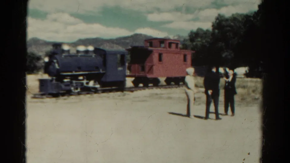 1967 Colorado frame with three visitors facing a stationary dark locomotive and red caboose under a partly cloudy sky.
