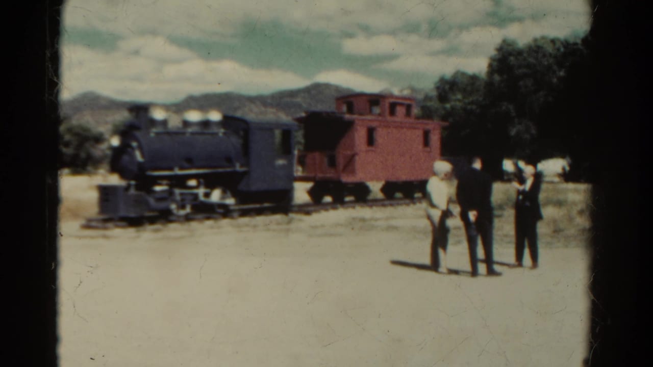 1967 Colorado 8mm frame showing a black steam locomotive, red caboose, and three visitors standing on open ground with mountains behind.