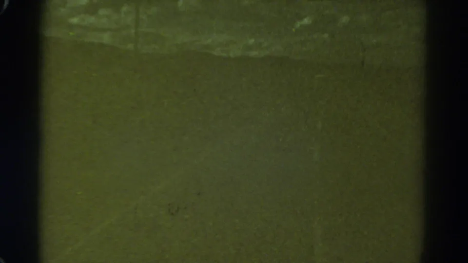1967 Colorado home-movie frame showing a dim nighttime road view with green tint, black film edges, grain, and a faint streak of light.