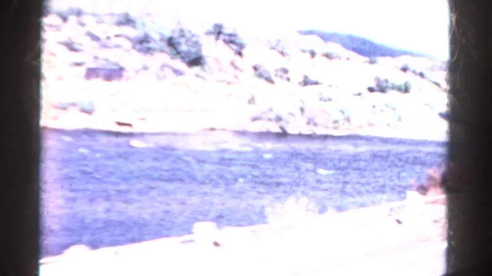 A bright 1967 Colorado 8mm view shows blue lake water below sunlit rocky hills and sparse vegetation.