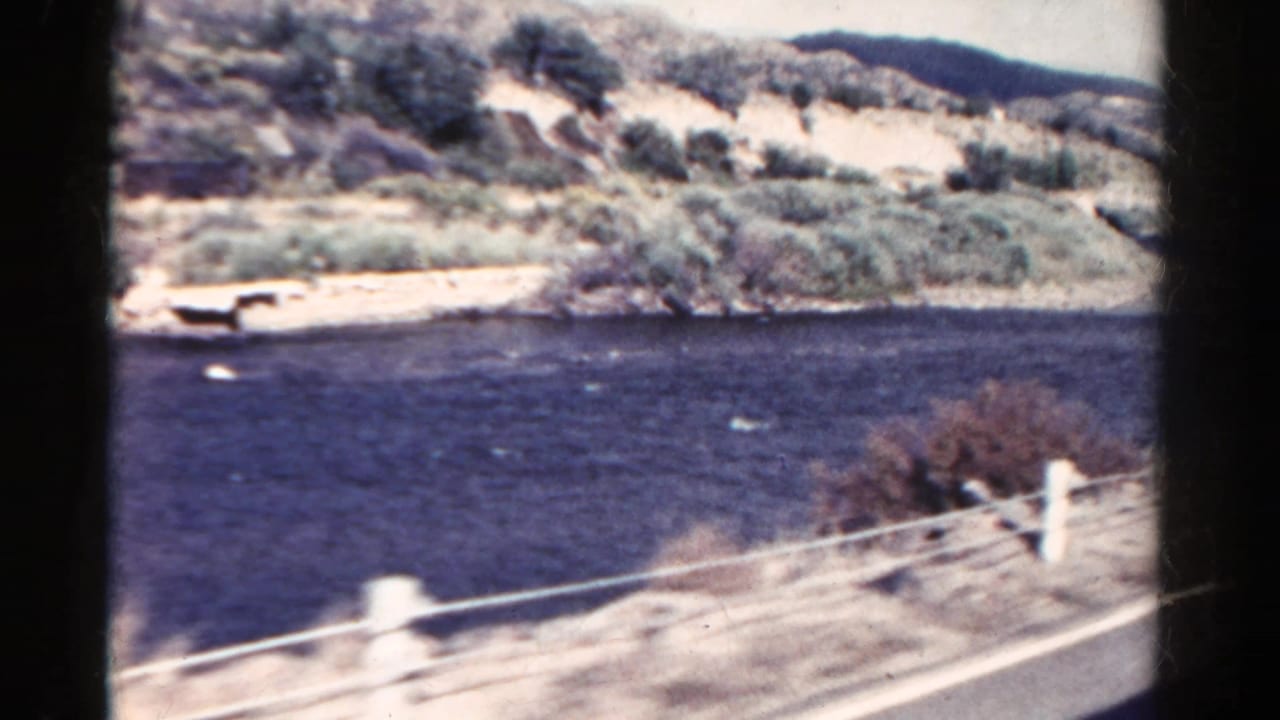 1967 Colorado 8mm frame showing a dark river, roadside railing, pale hills, and scrub vegetation from a moving car.