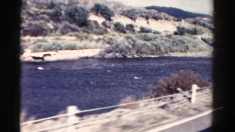 1967 Colorado 8mm frame showing a dark river, roadside railing, pale hills, and scrub vegetation from a moving car.