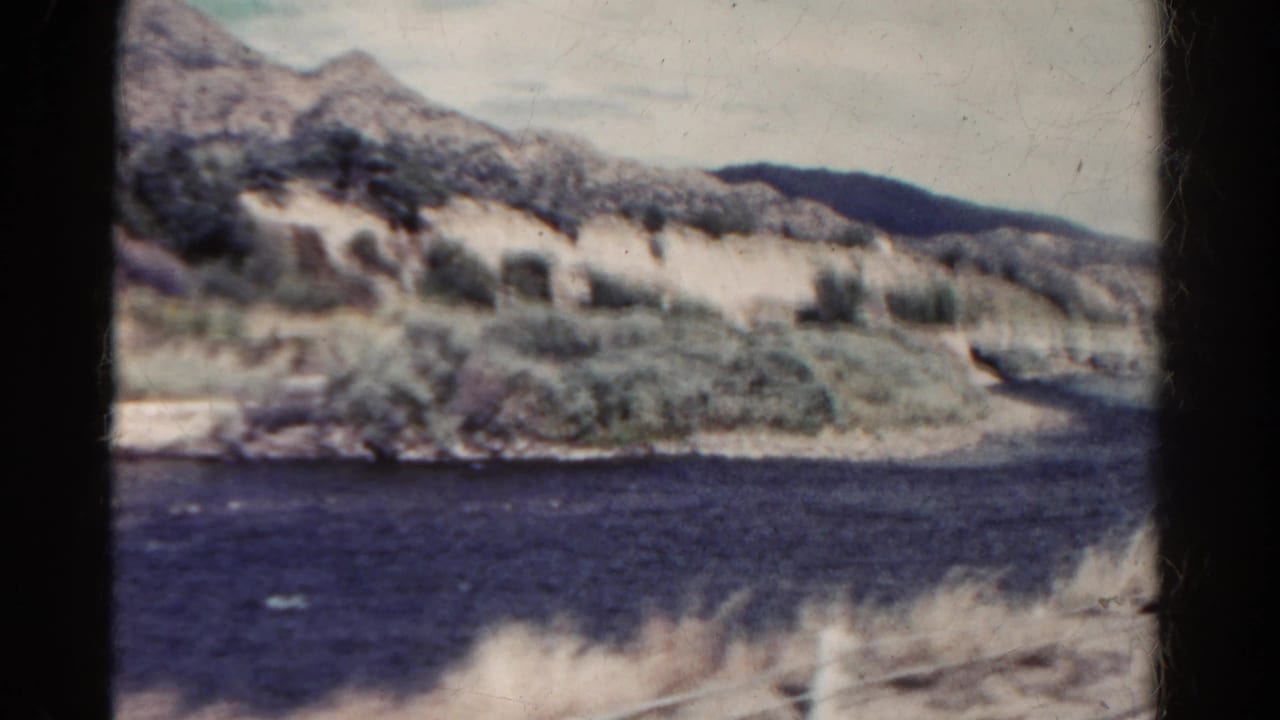 Colorado riverside landscape seen from a moving car, with arid hillside, shrubs, and dark water in a 1967 8mm frame.