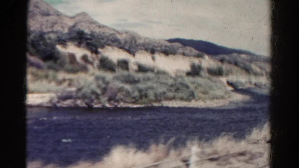 Colorado riverside landscape seen from a moving car, with arid hillside, shrubs, and dark water in a 1967 8mm frame.