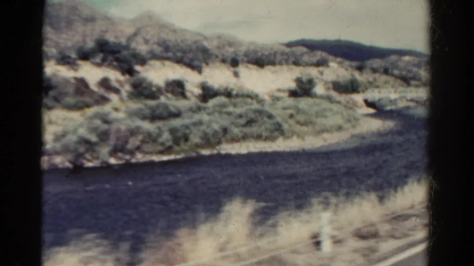 1967 Colorado home-movie frame of a winding river, pale hillside, sparse vegetation, and blurred roadside foreground.