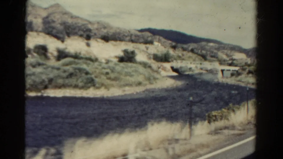 Colorado 8mm travel frame showing a river bend, dry hillside, sparse shrubs, roadside edge, and a small bridge-like structure.