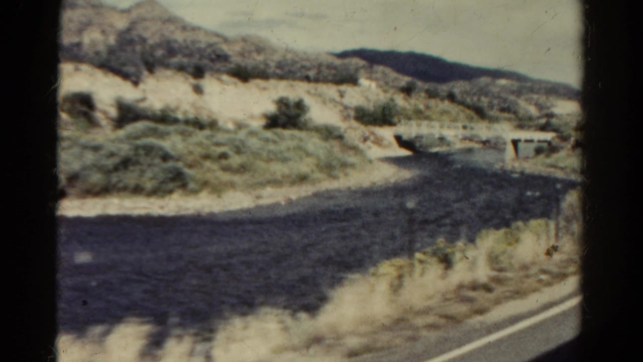 1967 Colorado 8mm frame showing a paved road, dry wash, low bridge, shrubs, and rocky hills from a moving-car viewpoint.