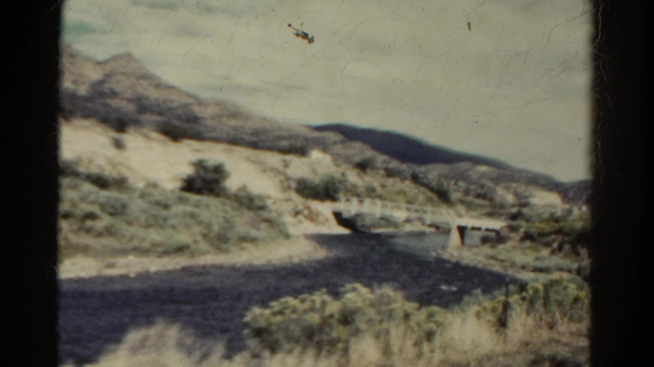 Vintage Colorado travel frame with a dry riverbed, small concrete bridge, scrub vegetation, and pale hills beyond the road.