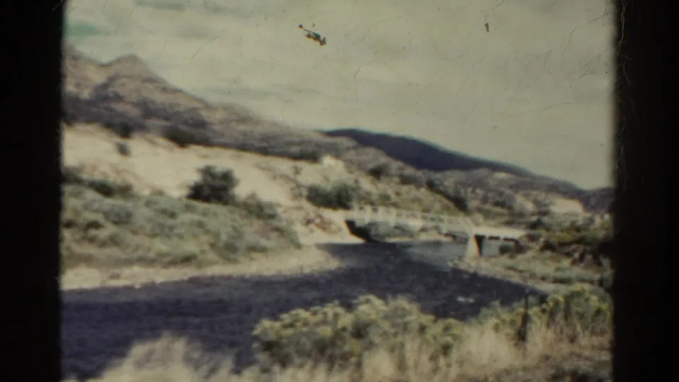 Vintage Colorado travel frame with a dry riverbed, small concrete bridge, scrub vegetation, and pale hills beyond the road.