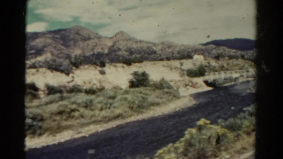 1967 home-movie frame from Colorado showing scrub-covered roadside, rocky hills, and part of a small bridge near a dark road.