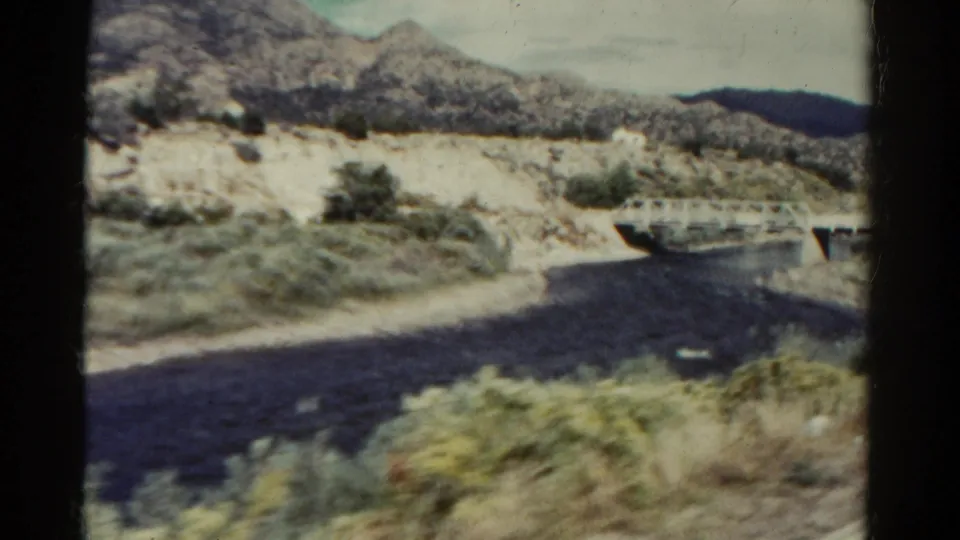 Vintage 8mm Colorado road scene with a dark paved curve, small concrete bridge, dry wash, roadside plants, and rugged hills.